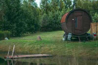 a large wooden barrel sitting on the grass next to a body of water at Šeimos Namelis Adomo Sodyboje prie ežero 35 km nuo Vilniaus šalia Dubingių in Dirmeičiai