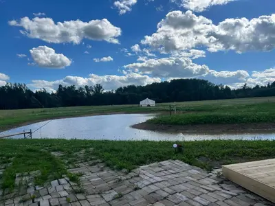 a pond in a field with a white house in the distance at Anos Kempingas, Troškūnai in Smėlynė
