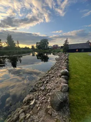 a river with a rock wall next to a field at Už Karvelius - Sodyba Biržų mieste in Biržai
